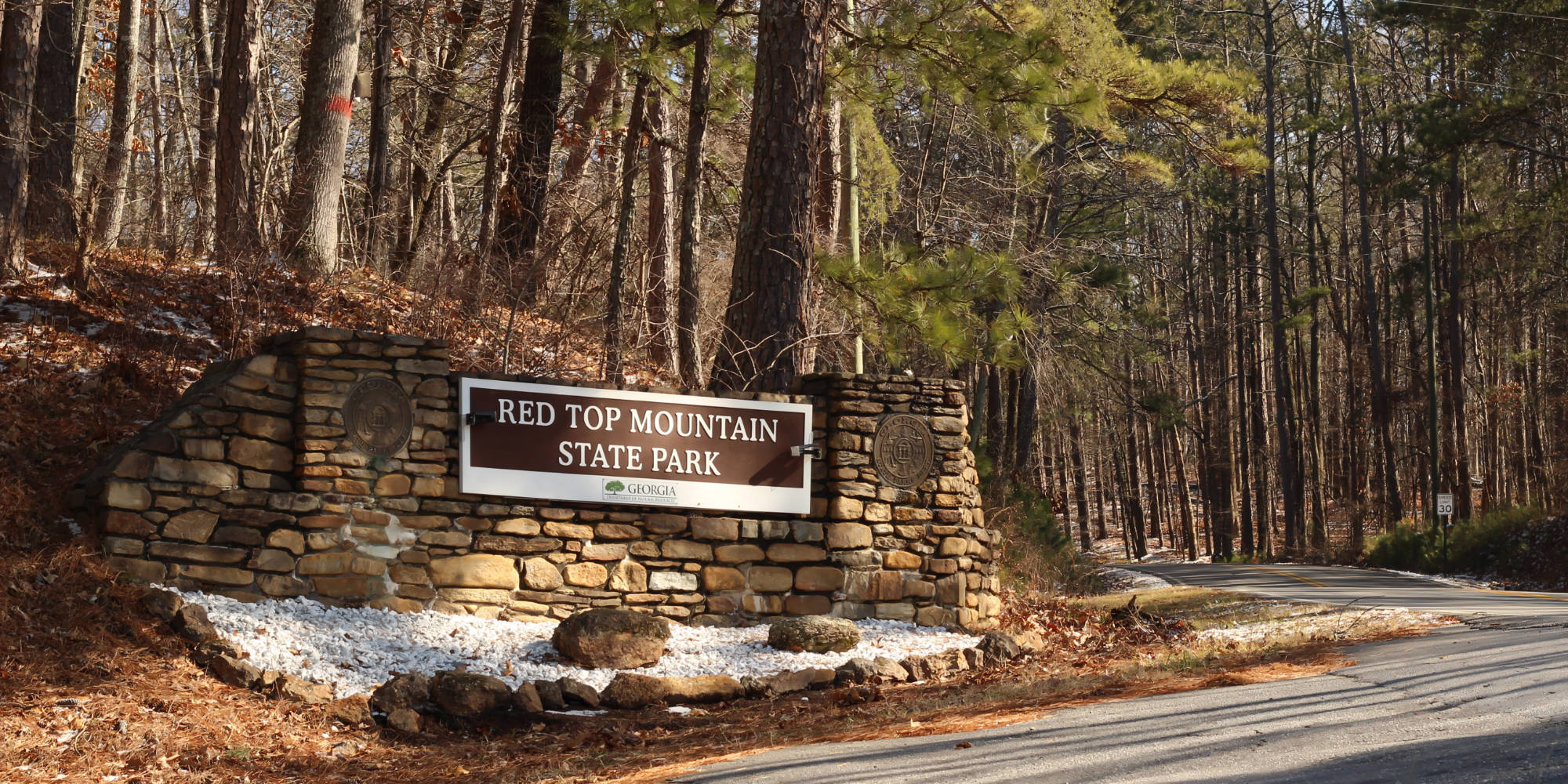 (a stone entrance sign on the side of the road for Red Top Mountain State Park, Georgia, surrounded by a pine tree forest)