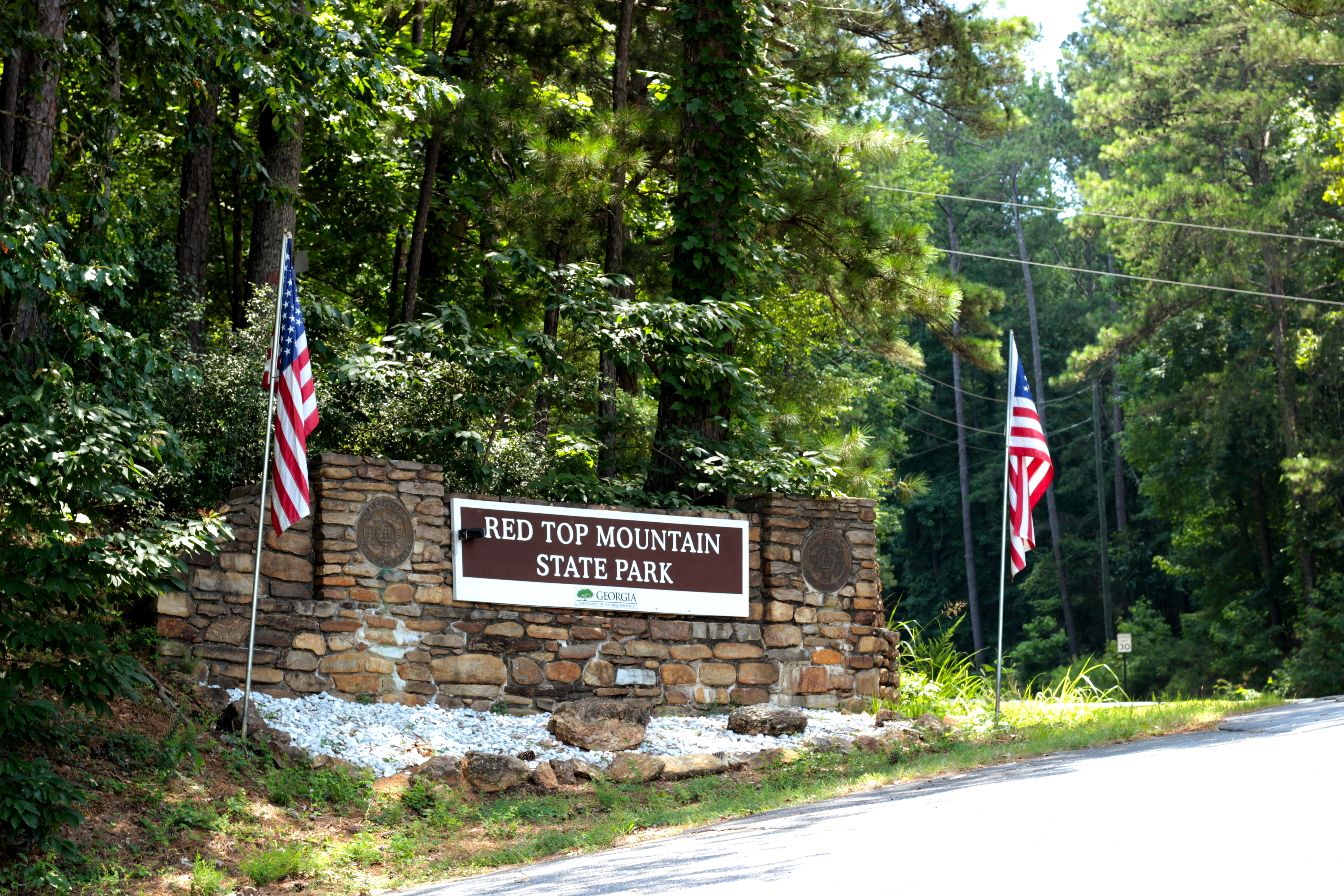 (a stone entrance sign on the side of the road for Red Top Mountain State Park, Georgia, surrounded by a forest. to the left and right of the sign there is a flag pole bearing a United States flag)